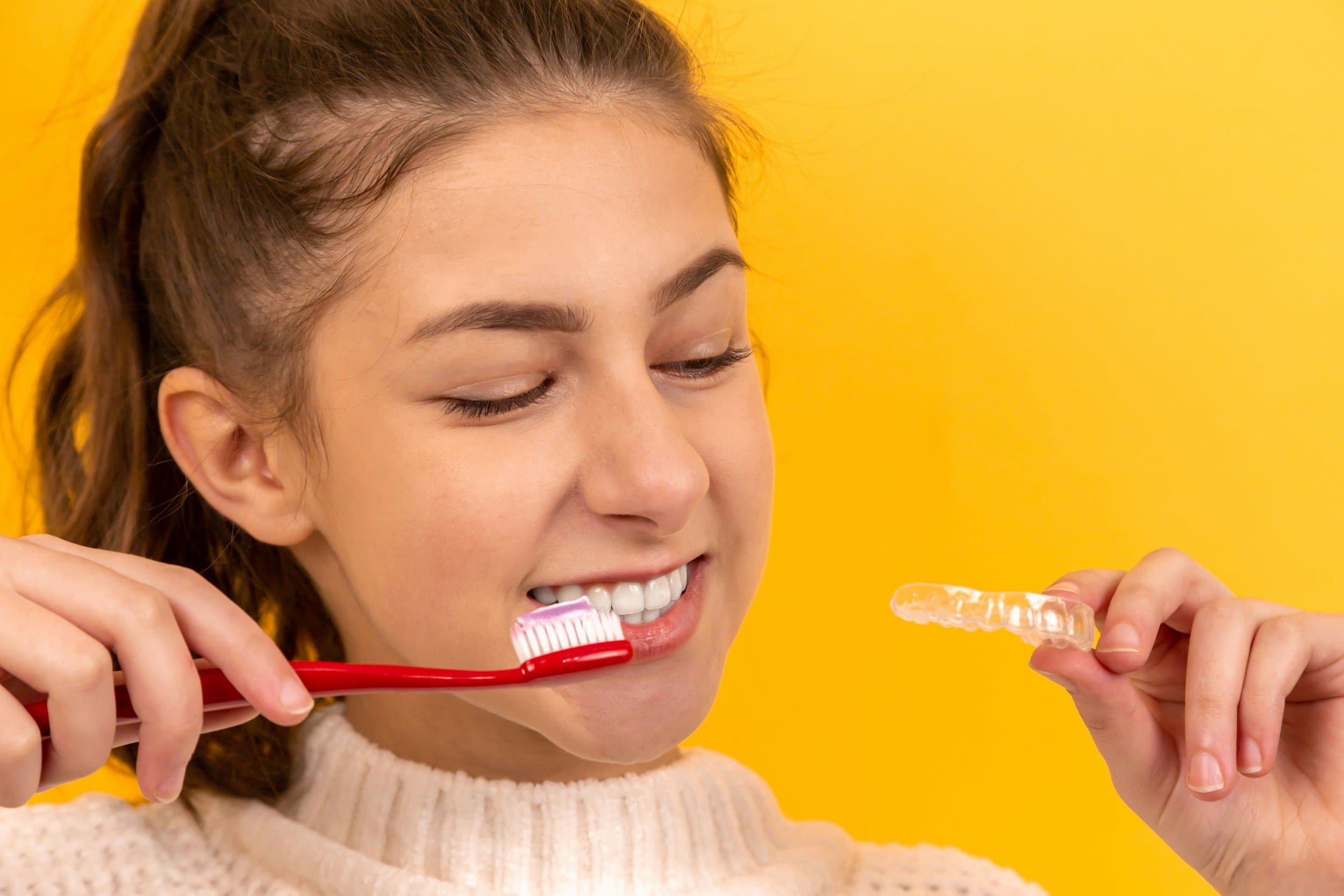 A young woman brushing her teeth with a clear retainer held up beside her against a yellow background, at Align Dental Surgery